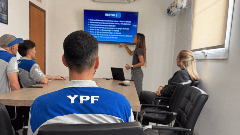 presentación o reunión de capacitación en una sala de juntas. Varias personas, algunas con uniforme de YPF, están mirando una pantalla donde una mujer expone